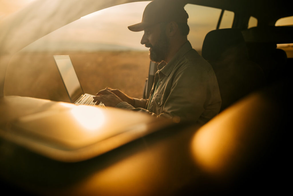 Man in vehicle working on laptop with field in the background